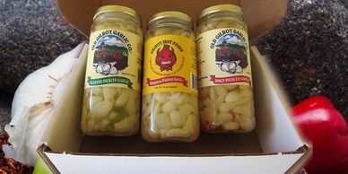 3 jars of pickled garlic in a white gift box on top of garlic, peppers and chili flakes