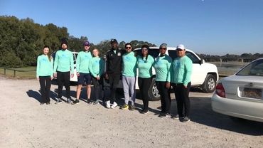 Group of people in turquoise shirts posing outdoors near parked cars.