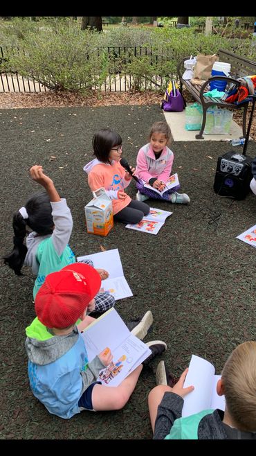 Children sitting outdoors, reading and engaging in a group activity.