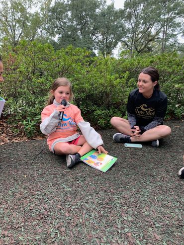 A young girl reads aloud outdoors while others listen attentively.