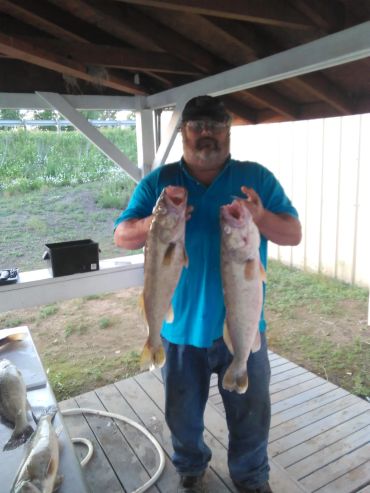 Man proudly holding two large fish under a wooden shelter.