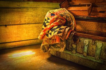 "Comfort."  An old quilt on the edge of an antique trunk catches sunlight through a window.