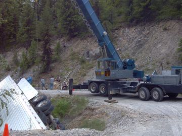 large blue crane attempts to lift a large white over turned semi from side of mountain cliff
